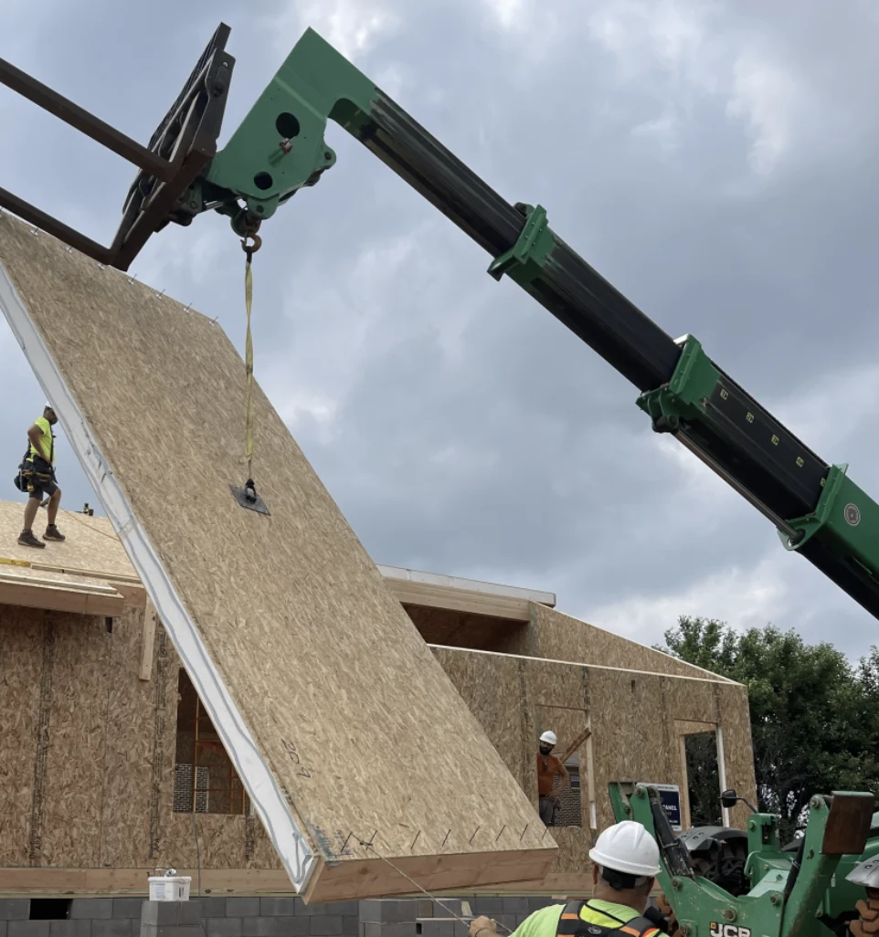 A crane positioning a large insulated roof panel above a residential building enclosure